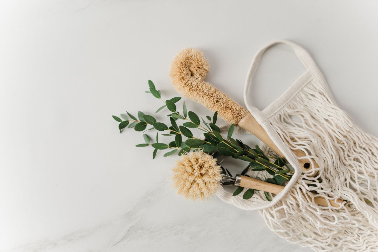 Brosses de nettoyage écologiques et branches d’eucalyptus dans un sac en filet réutilisable, posés sur une surface blanche.
